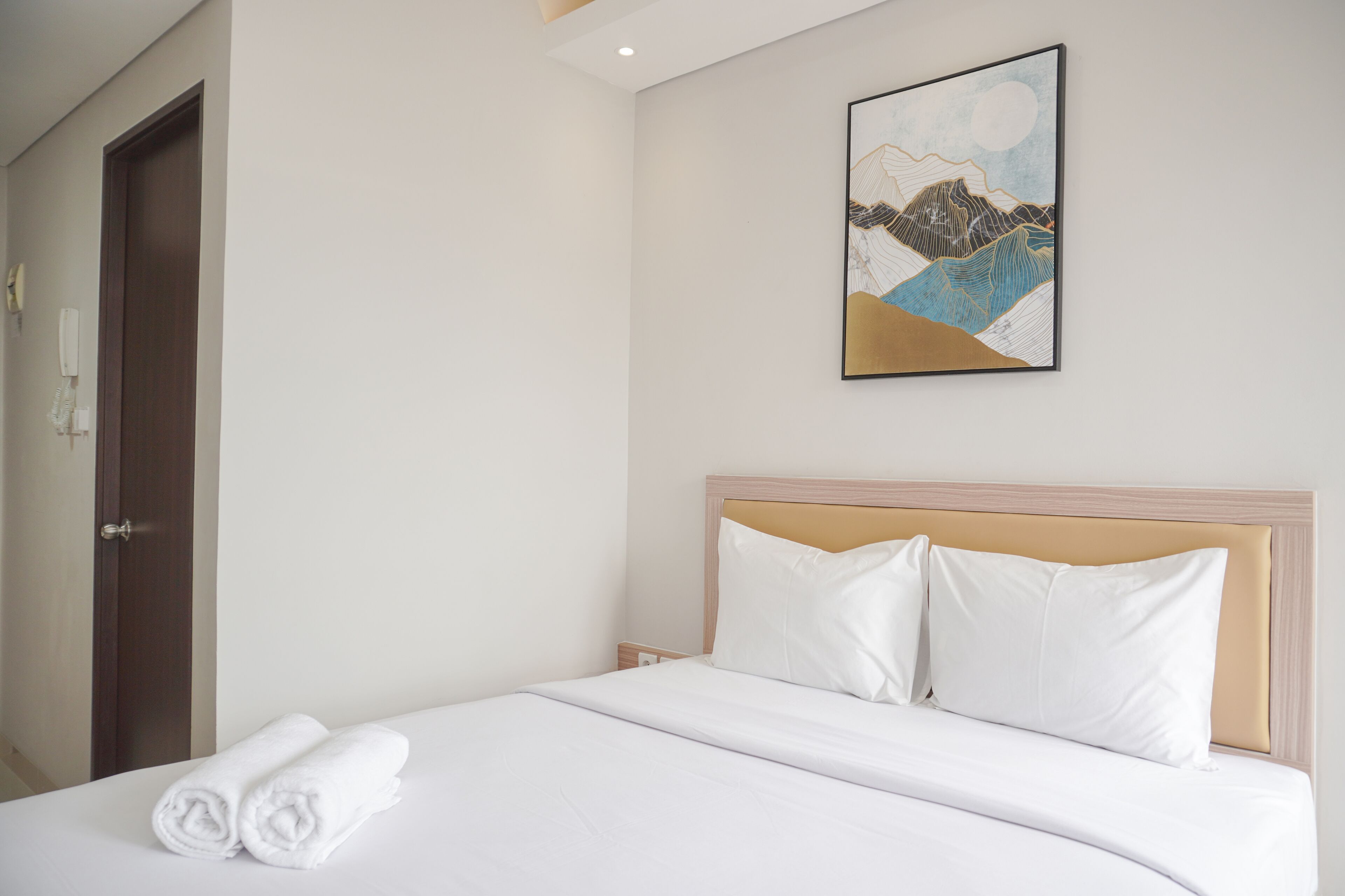 A neatly made bed with white linens and pillows, a wooden headboard, and a framed artwork on the wall.