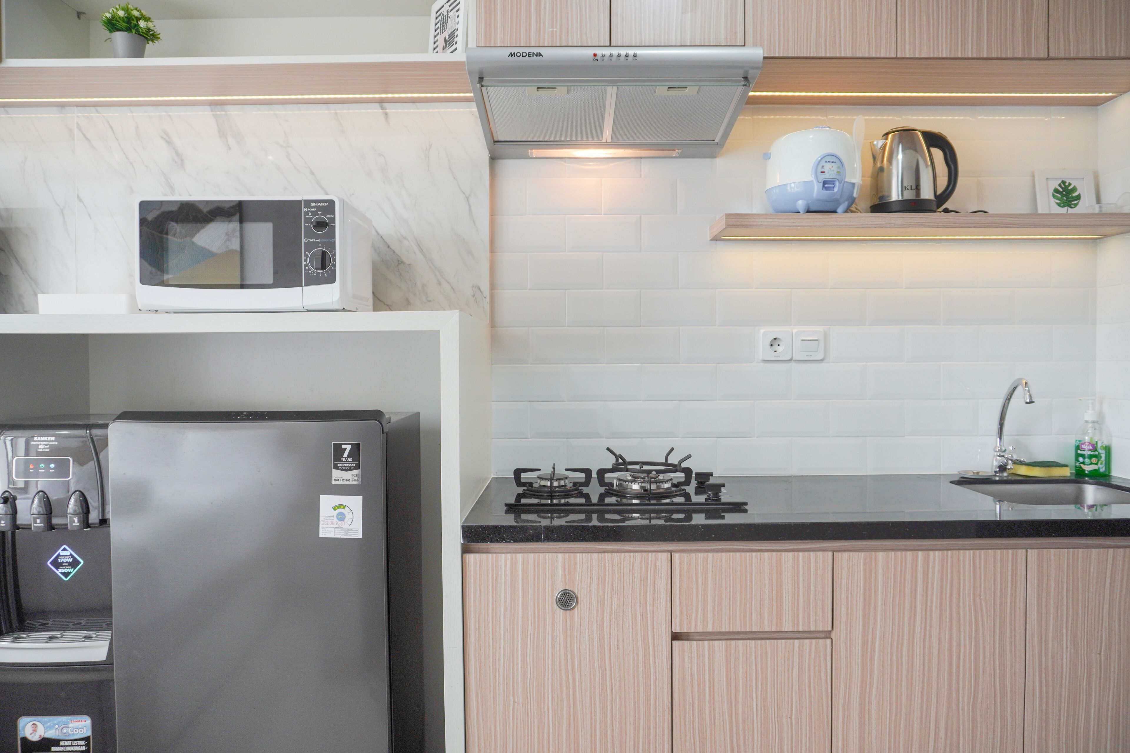 A kitchen with a microwave, refrigerator, stove, and a shelf with a kettle and a plant.