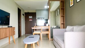 A modern living room with a grey sofa, a small round table, and a television on a stand. There is a wooden cabinet and a kitchen area visible in the background.