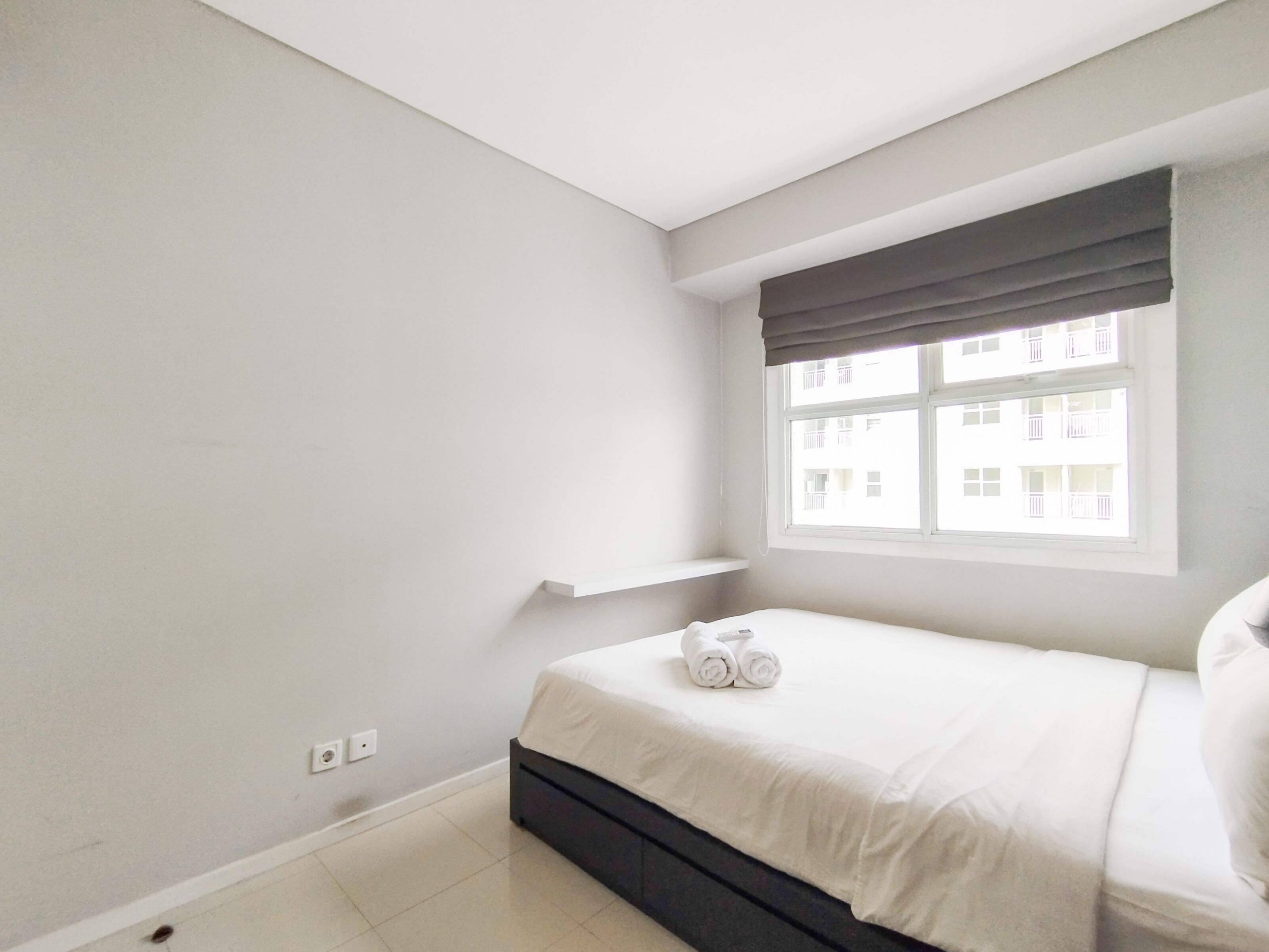 A bedroom with a bed, a window with a view of buildings, and a white wall.