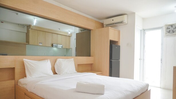 A modern bedroom with a wooden bed frame, white bedding, and a built-in wardrobe. - Good Deal And Homey Studio Bassura City Apartment (Jakarta)