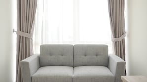 A room with a light grey upholstered sofa positioned in front of a window with sheer curtains.