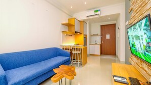 A modern living room with a blue sofa, a wooden coffee table, a TV, and a kitchen area in the background. - Best And Stylish 2Br At Signature Park Grande Apartment (Jakarta)