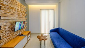 A modern living room with a wooden wall panel, a blue sofa, a wooden coffee table, and a TV mounted on the wall. - Best And Stylish 2Br At Signature Park Grande Apartment (Jakarta)