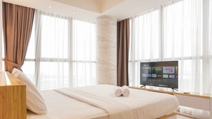 A modern hotel room with a large bed, a flat-screen TV, and large windows with sheer curtains. - Modern Look And Nice 2Br Gold Coast Apartment Near Pik (Jakarta)