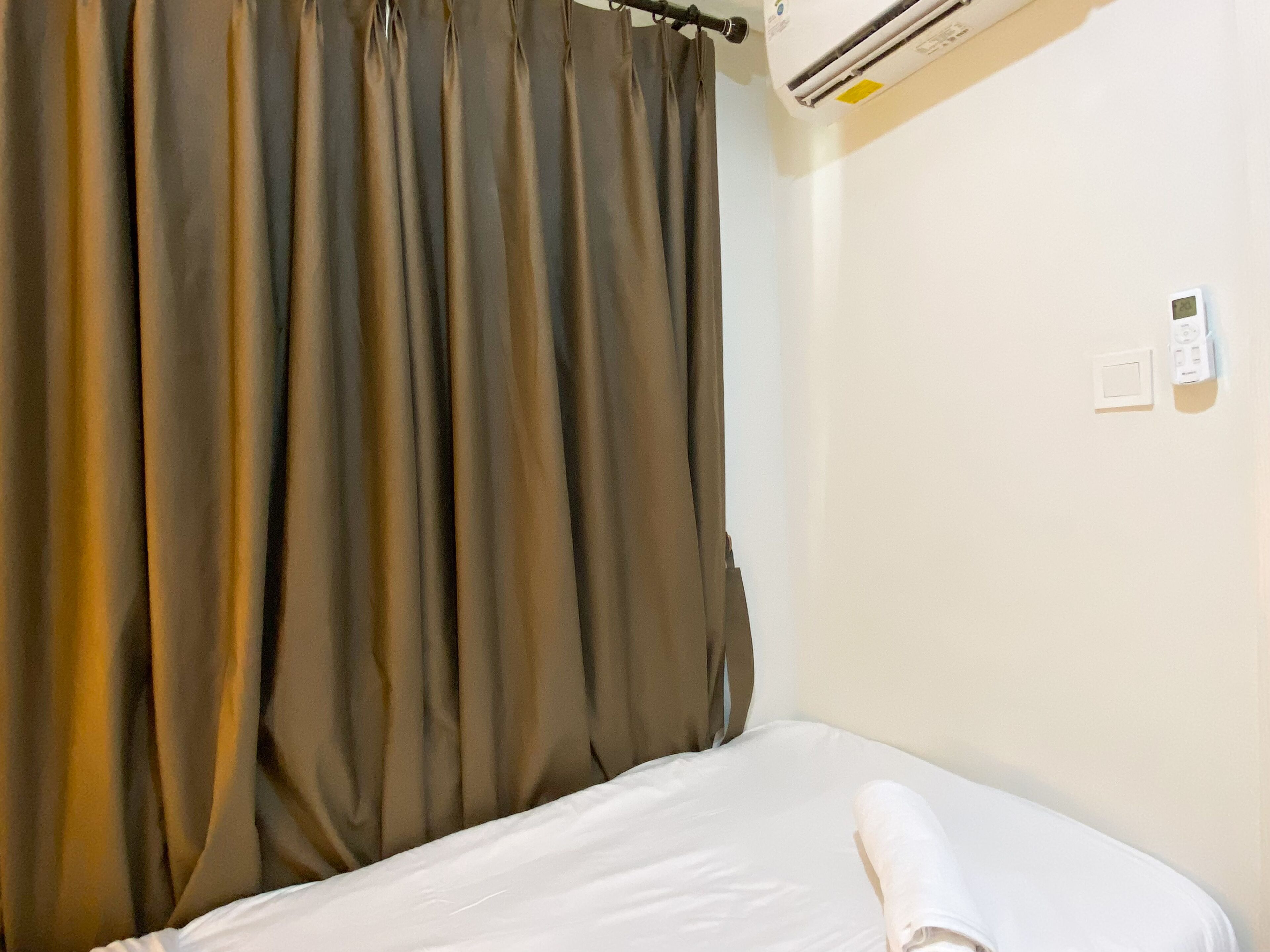 Una cama individual con ropa blanca, una cortina marrón, un equipo de aire acondicionado y un termostato en la pared.