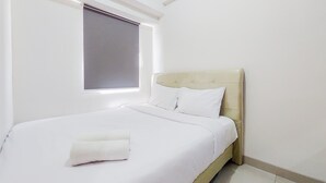 Cama bem arrumada com lençóis brancos e uma toalha dobrada, cabeceira estofada em bege e janela com persiana parcialmente abaixada.