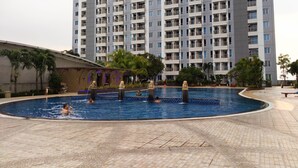 A swimming pool with a tiled deck, surrounded by palm trees and a high-rise building in the background. - Homey Studio At Orchard Supermall Mansion Apartment (Surabaya)