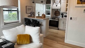 A modern living room with a sofa, a coffee table, and a black and white rug. A kitchen is visible in the background with a stove, microwave, and cabinets. - Black Brindle Cottage, Downtown Ft. Myers: Walk to Best Eats and Shops! (Fort Myers)