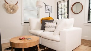 Un canapé blanc agrémenté d’un plaid jaune moutarde, une table ronde en bois sur laquelle sont posés des livres et une télécommande, et un tapis à longues mèches.