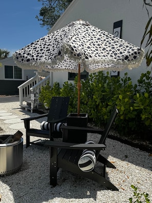 A patio with a patterned umbrella, a chair, a small table, and a trash can.