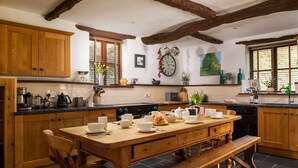 A kitchen with wooden cabinets, a large wooden table, and a bench. There is a clock on the wall and a window with plants on the windowsill.