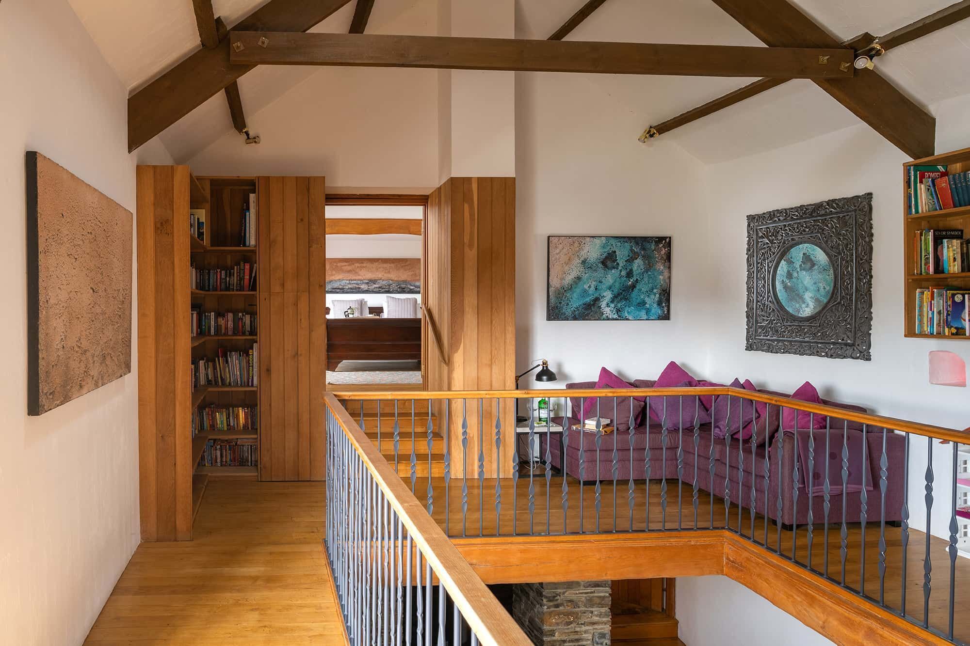 A spacious loft with exposed wooden beams, a wooden staircase, a purple sofa, and a bookshelf filled with books.