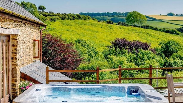Un jacuzzi en una terraza de madera con un edificio de piedra y vistas a colinas verdes.
