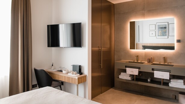 A modern hotel room with a bed, a desk with a chair, a flat-screen TV, a large mirror, and a dual sink vanity. - Boutique HOH (Zagreb)