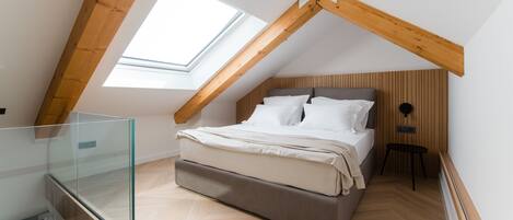 Un dormitorio con techo inclinado, cama con ropa de cama blanca, cabecera de madera y barandilla de vidrio.