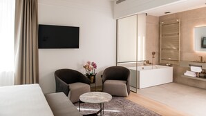 Una habitación de hotel moderna con una cama grande, dos sillones, una mesita redonda, un televisor de pantalla plana, un espejo grande, una bañera y un tocador con espejo.
