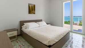 Une chambre à coucher comprenant un lit, une table de chevet, un tiroir, un tableau et une vue sur la mer, visible à travers une porte coulissante.