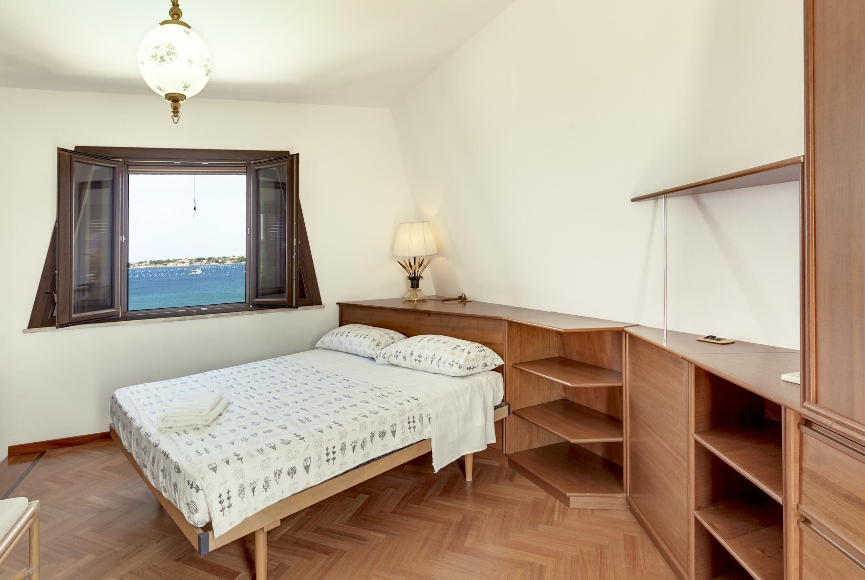 Een slaapkamer met een bed, een bureau, een lamp en uitzicht op de zee door een raam.