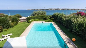 Une piscine rectangulaire à l’eau d’un bleu cristallin, entourée d’une végétation luxuriante et bien entretenue, et offrant une vue sur la mer.