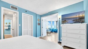 A bedroom with a large bed, a dresser, and a view of the living area. - 🏖️Beach Front with Great Views - 1BR Sunbird 606W🏖️ (Panama City Beach)
