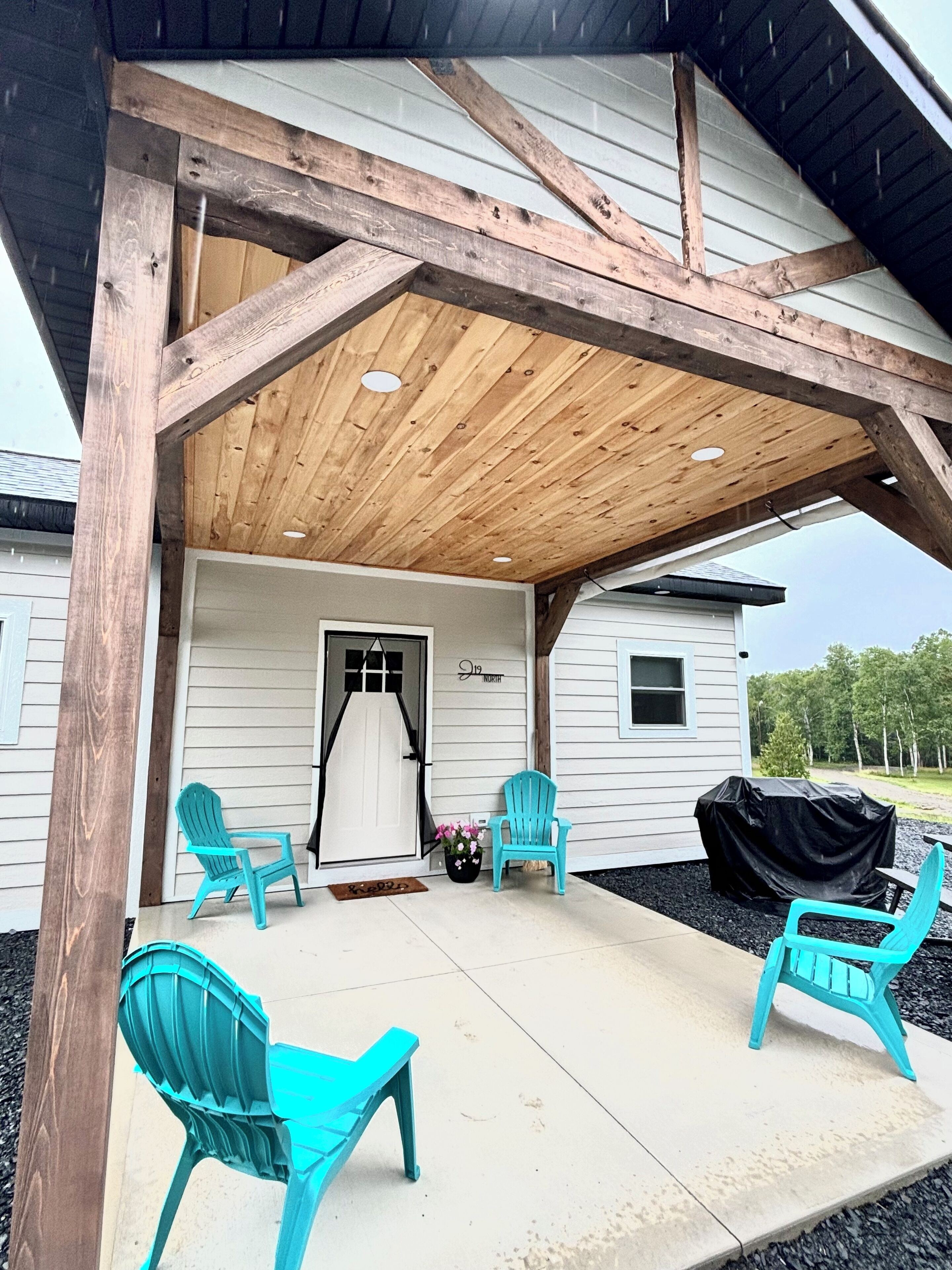 A covered porch with wooden beams and a concrete floor, featuring two turquoise chairs and a grill.