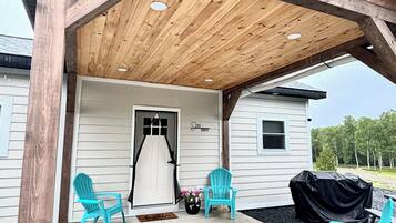 A covered porch with wooden beams and a concrete floor, featuring two turquoise chairs and a grill.