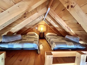 A small attic bedroom with two beds, wooden beams, and a single wall-mounted lamp.