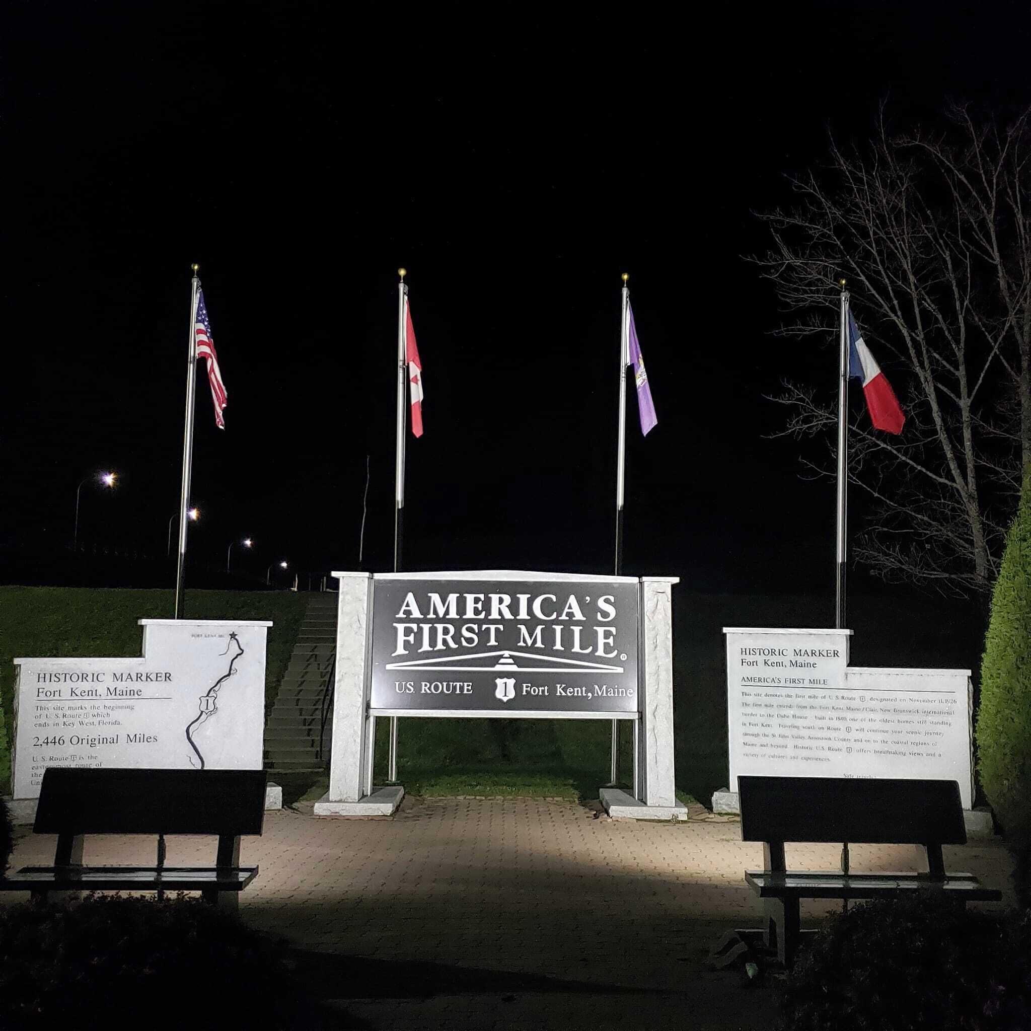 A nighttime view of America's First Mile sign with three flags on flagpoles, a bench, and a historical marker plaque.