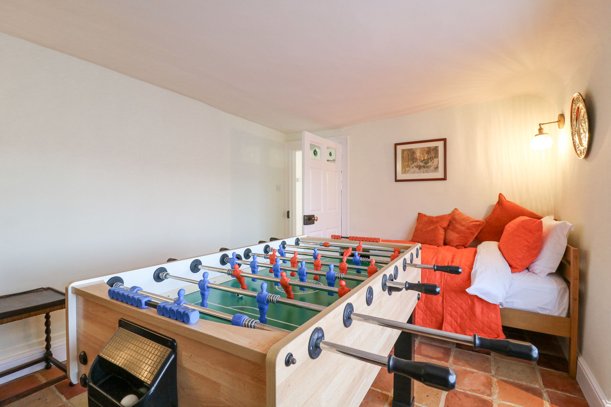 A room with a foosball table, a bed with orange pillows, and a wall-mounted light fixture.