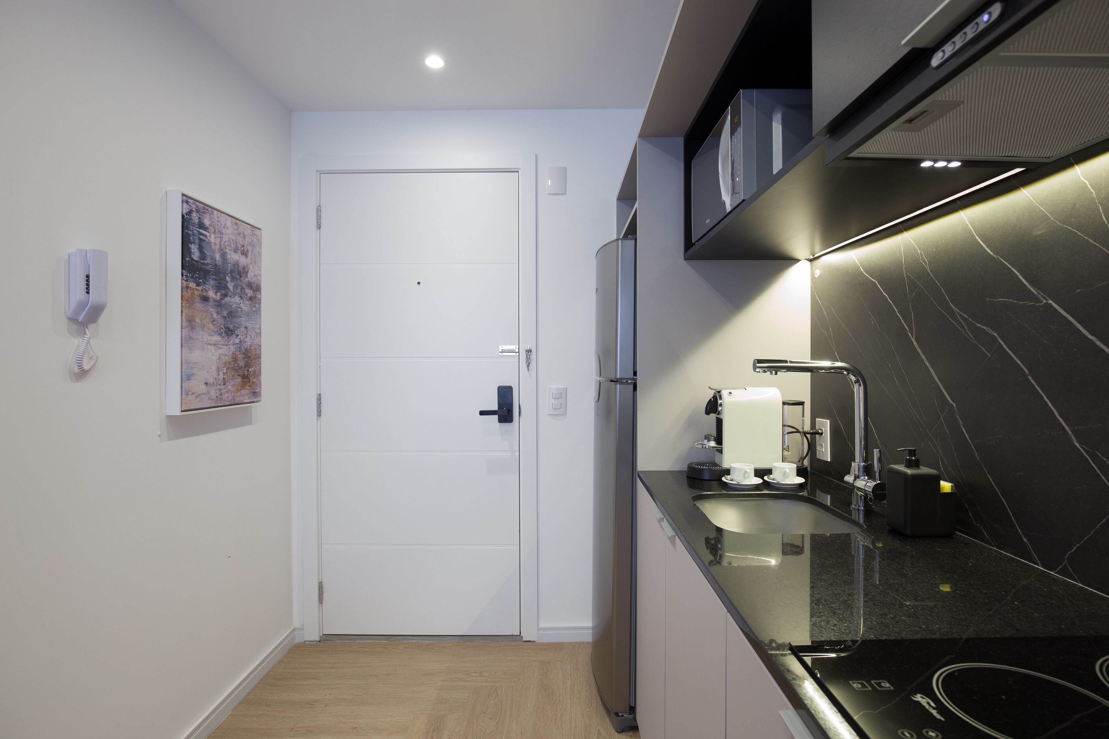 Una cocina compacta con una puerta blanca, un refrigerador, una encimera negra y un teléfono instalado en la pared.