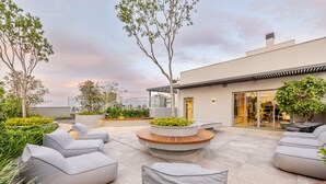 Un patio al aire libre con mobiliario moderno, una terraza de madera y un edificio con grandes ventanales y una puerta.