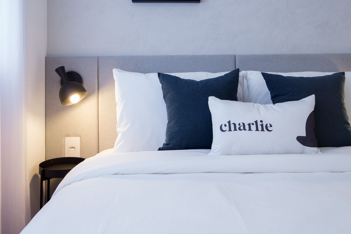 Una cama bien hecha con almohadas blancas y azul marino, una de ellas con el nombre 'charlie' bordado, y un cuadro enmarcado sobre la cama.