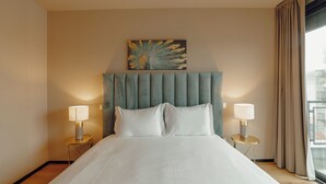 Een net opgemaakt bed met witte lakens, twee bedlampen en een schilderij boven het hoofdeinde.
