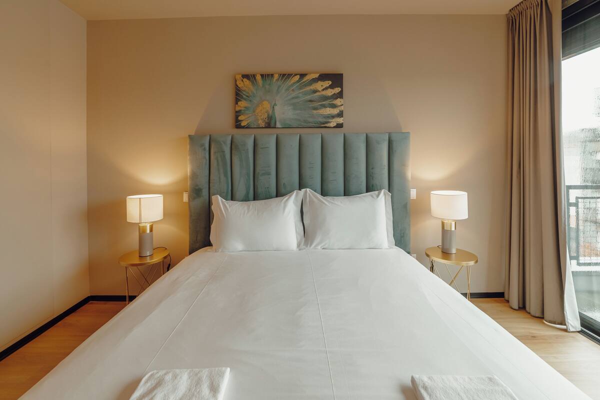 A neatly made bed with white linens, two bedside lamps, and a painting above the headboard.