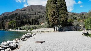 Una spiaggia di ciottoli con un grande albero, una panchina e una vista libera sull'acqua e le colline sullo sfondo.
