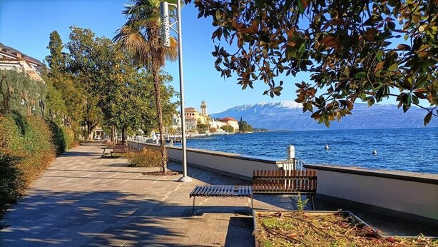 Una passeggiata lastricata sul lungomare, con panchine, un corrimano e vista sul mare e sulle montagne.