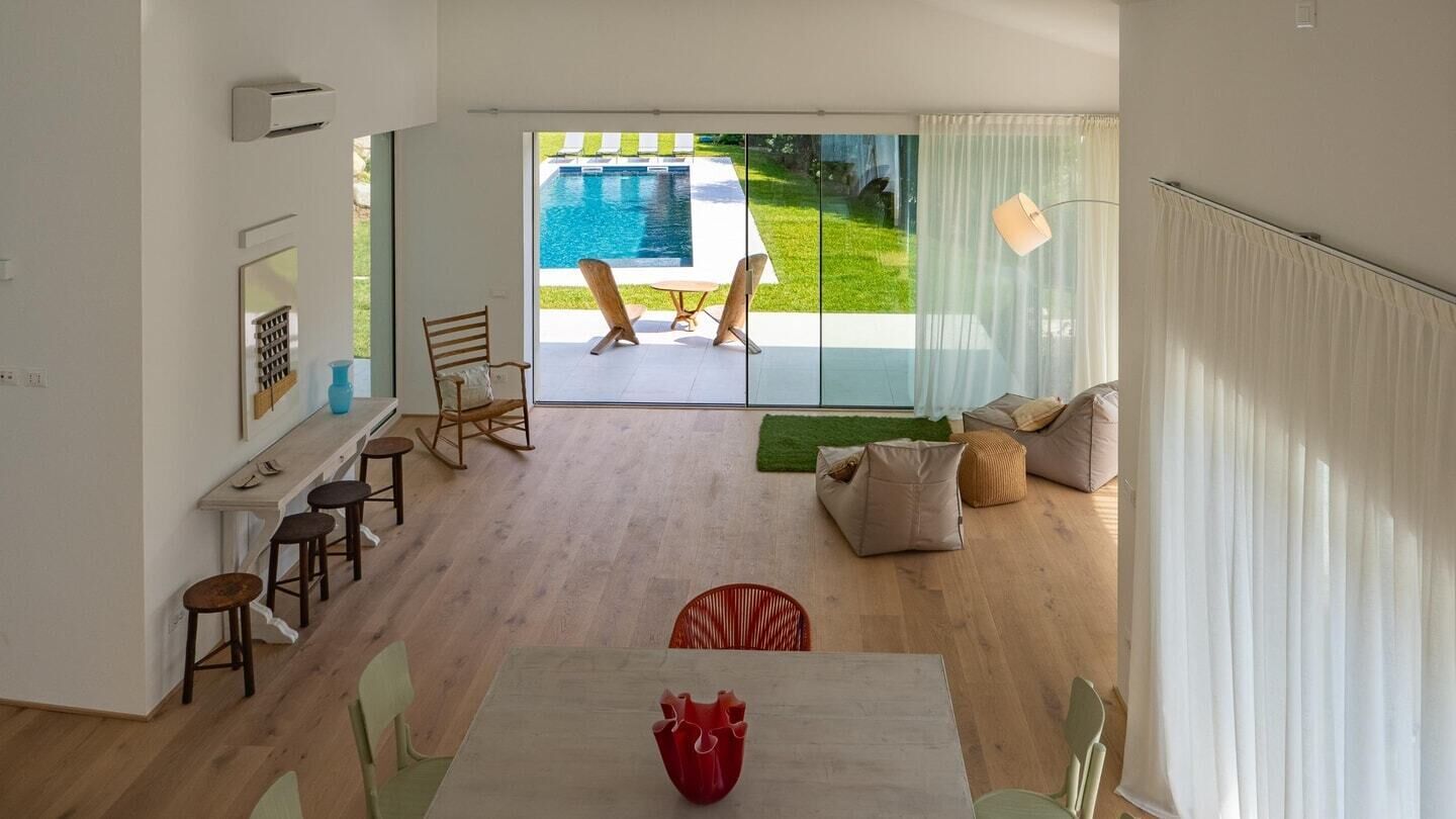 A spacious living area with a large sliding glass door leading to a pool, a dining table, and a seating area with cushions.