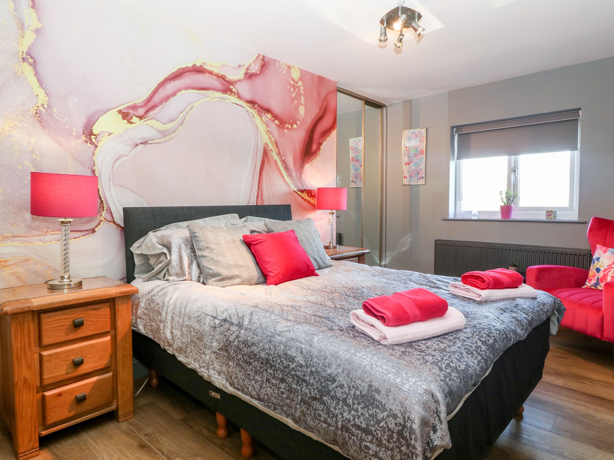 A bedroom with a bed, two bedside tables, a nightstand, a chair, a window, and a radiator.