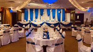 Un salón de banquetes con mesas redondas dispuestas para un evento formal, cubiertas con manteles azul y blanco, y una gran decoración floral azul colgando del techo.