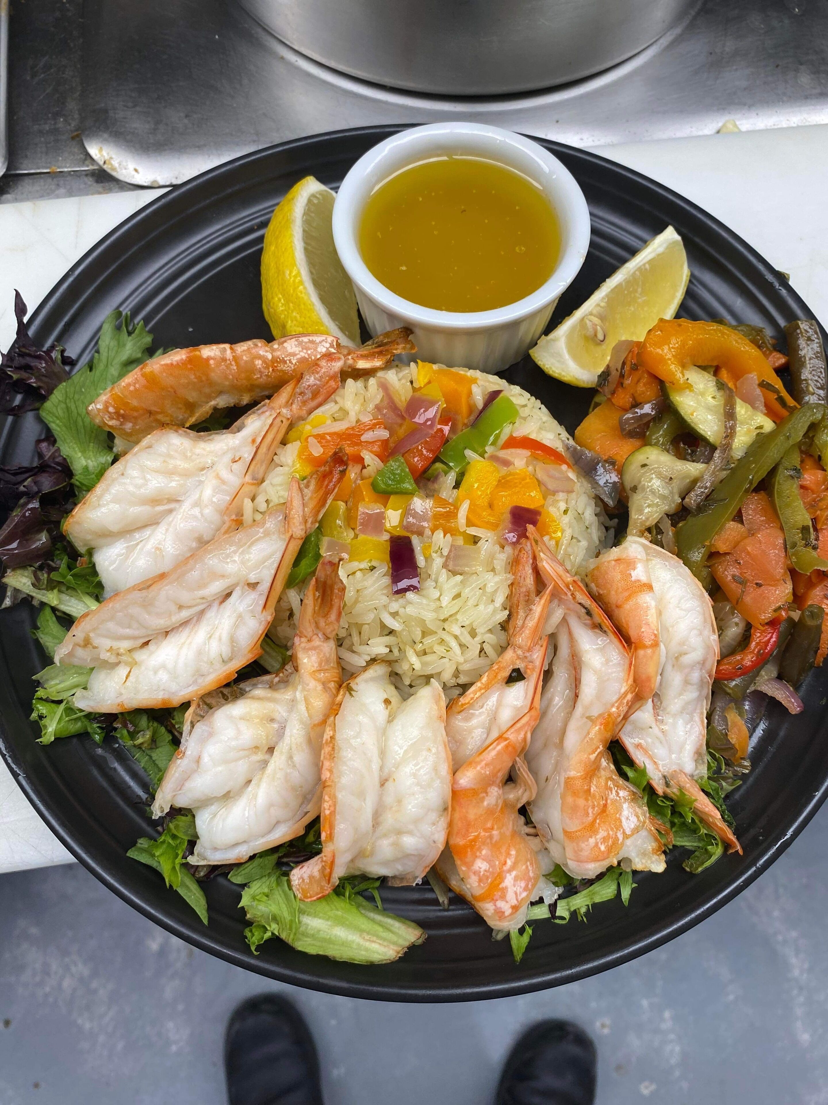 En tallerken mad med rejer, ris og grøntsager, samt en lille skål sauce og skiver af citron.