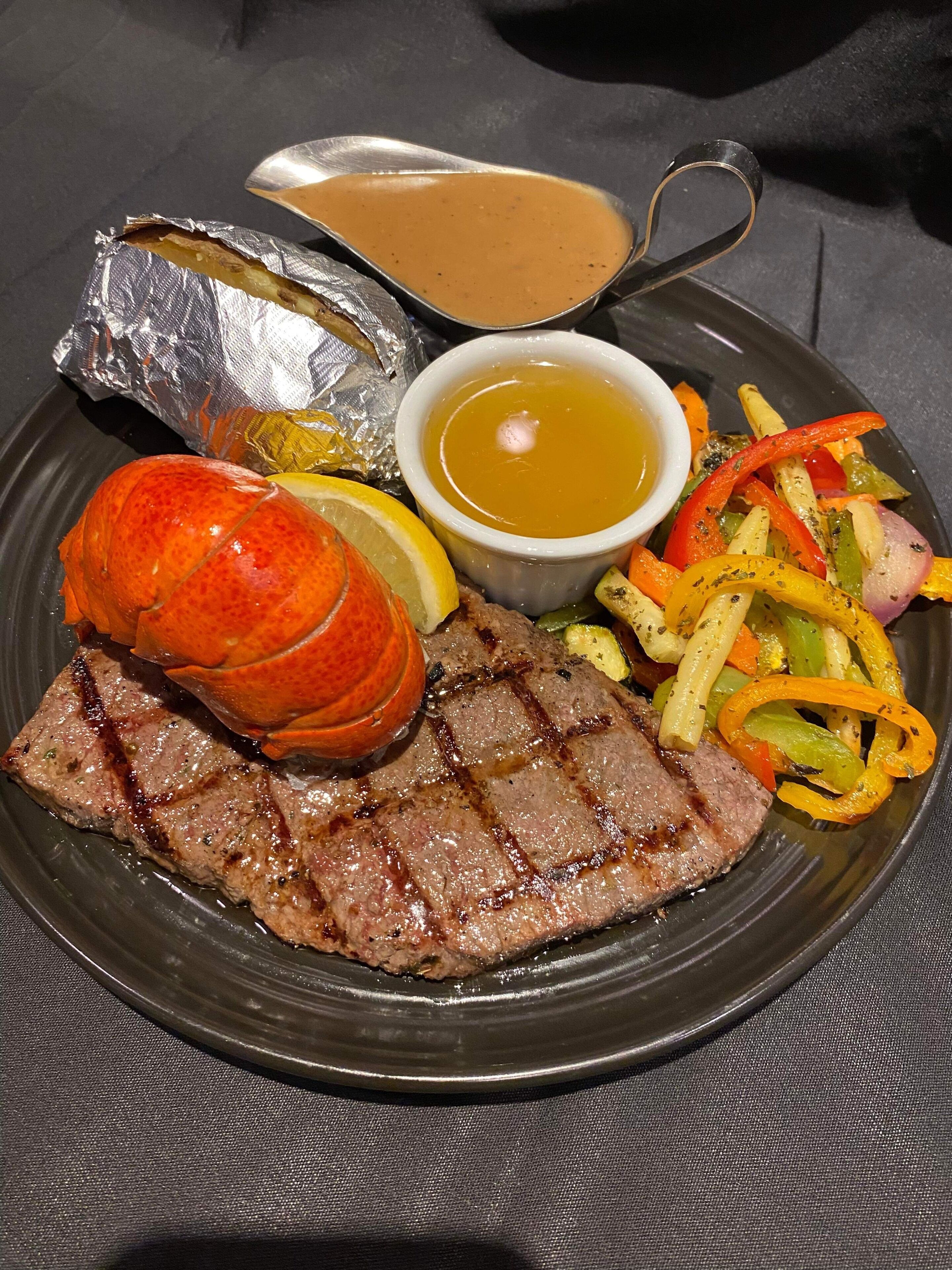 A plate with grilled steak, a lobster, roasted potatoes, and a side of mixed vegetables.