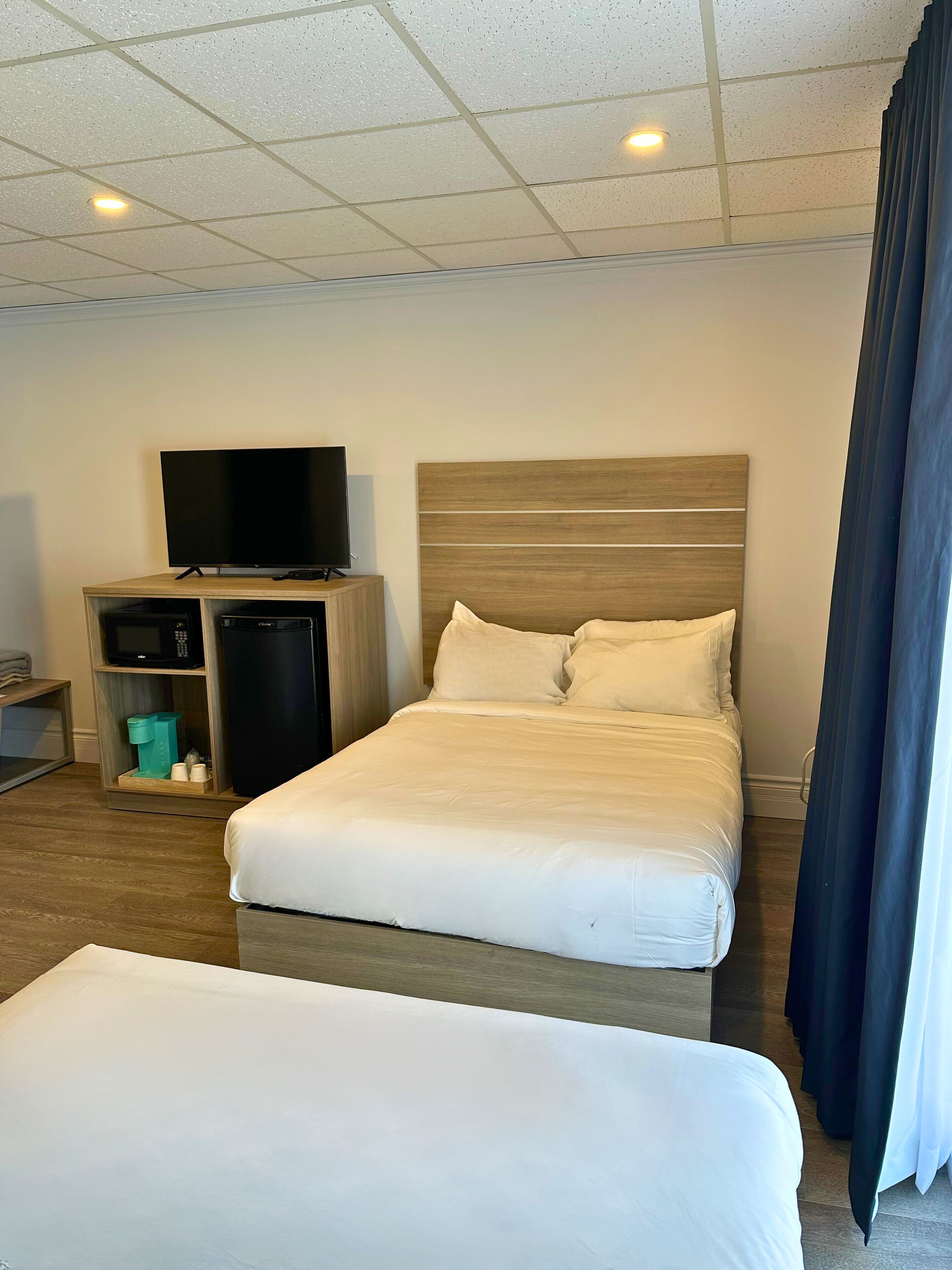 A hotel room with a large bed, a TV on a wooden stand, a small kitchenette, and a window with curtains.