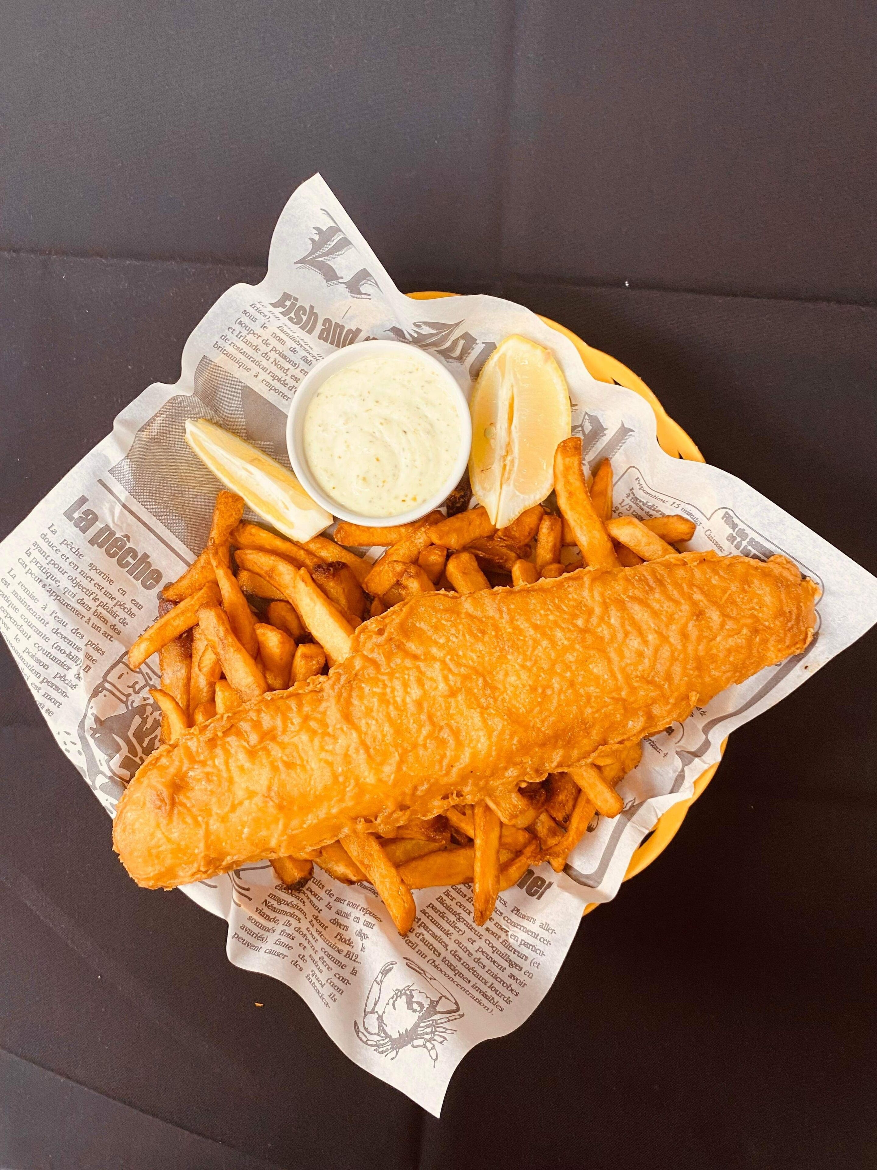 Una cesta de pescado con papas fritas, cuñas de limón y un recipiente pequeño de salsa.