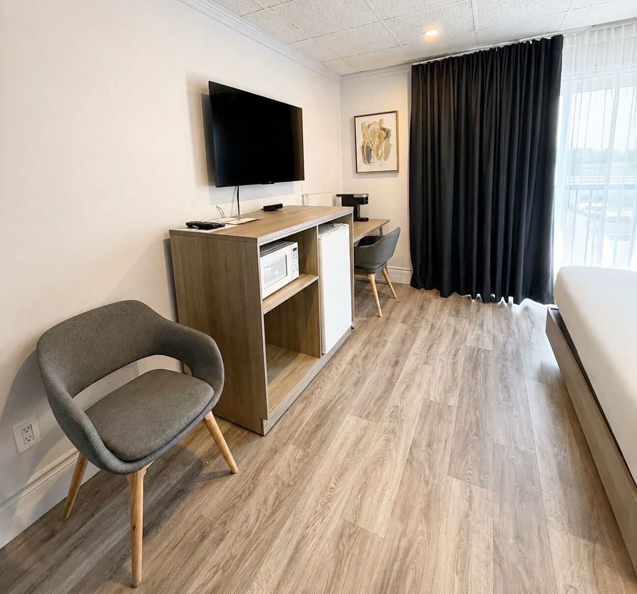 A hotel room with a bed, a desk with a flat-screen TV, a microwave, a chair, and a large window with curtains.