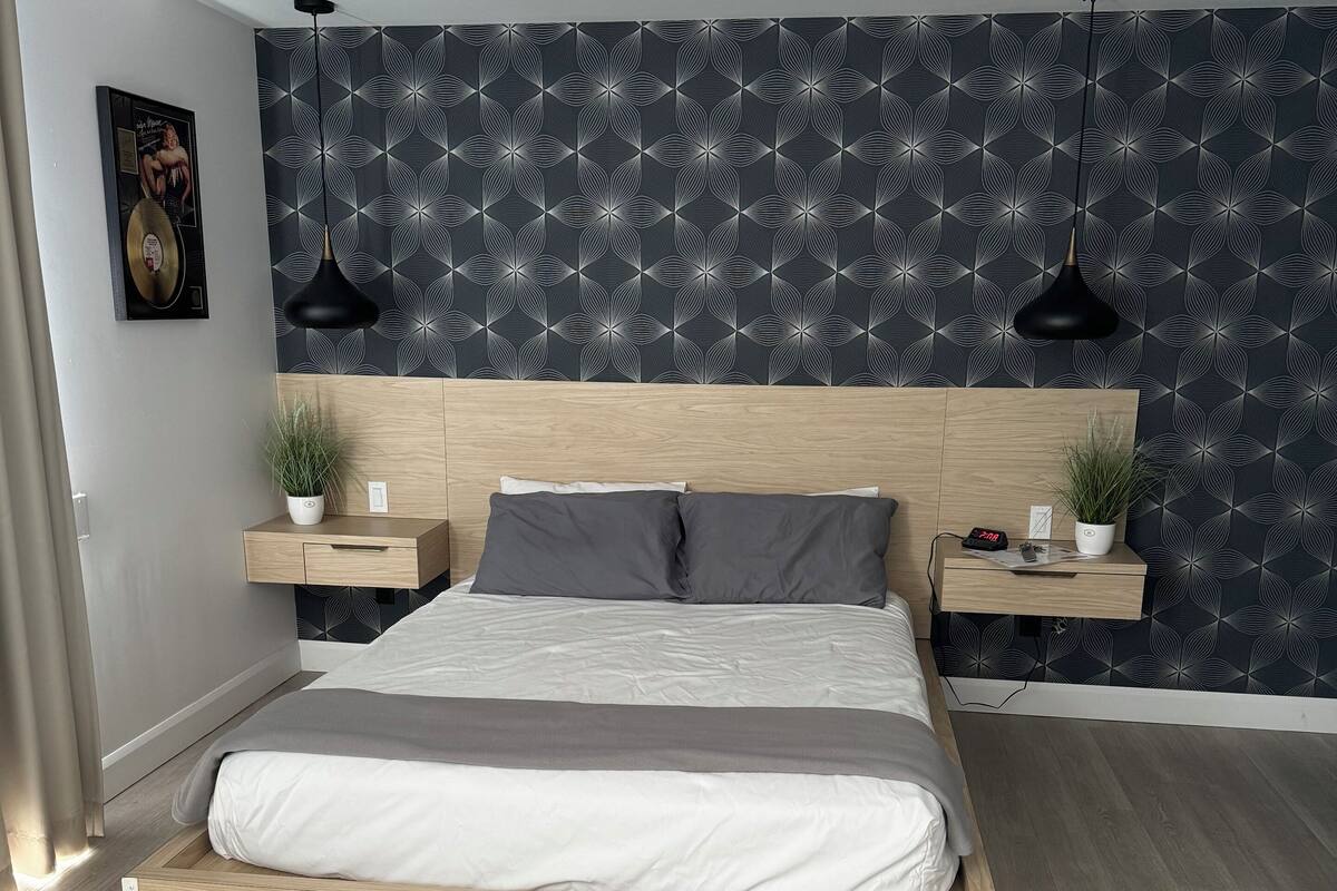 Un dormitorio moderno con una cama de madera, ropa de cama gris, un papel tapiz estampado y dos mesitas de noche con plantas.