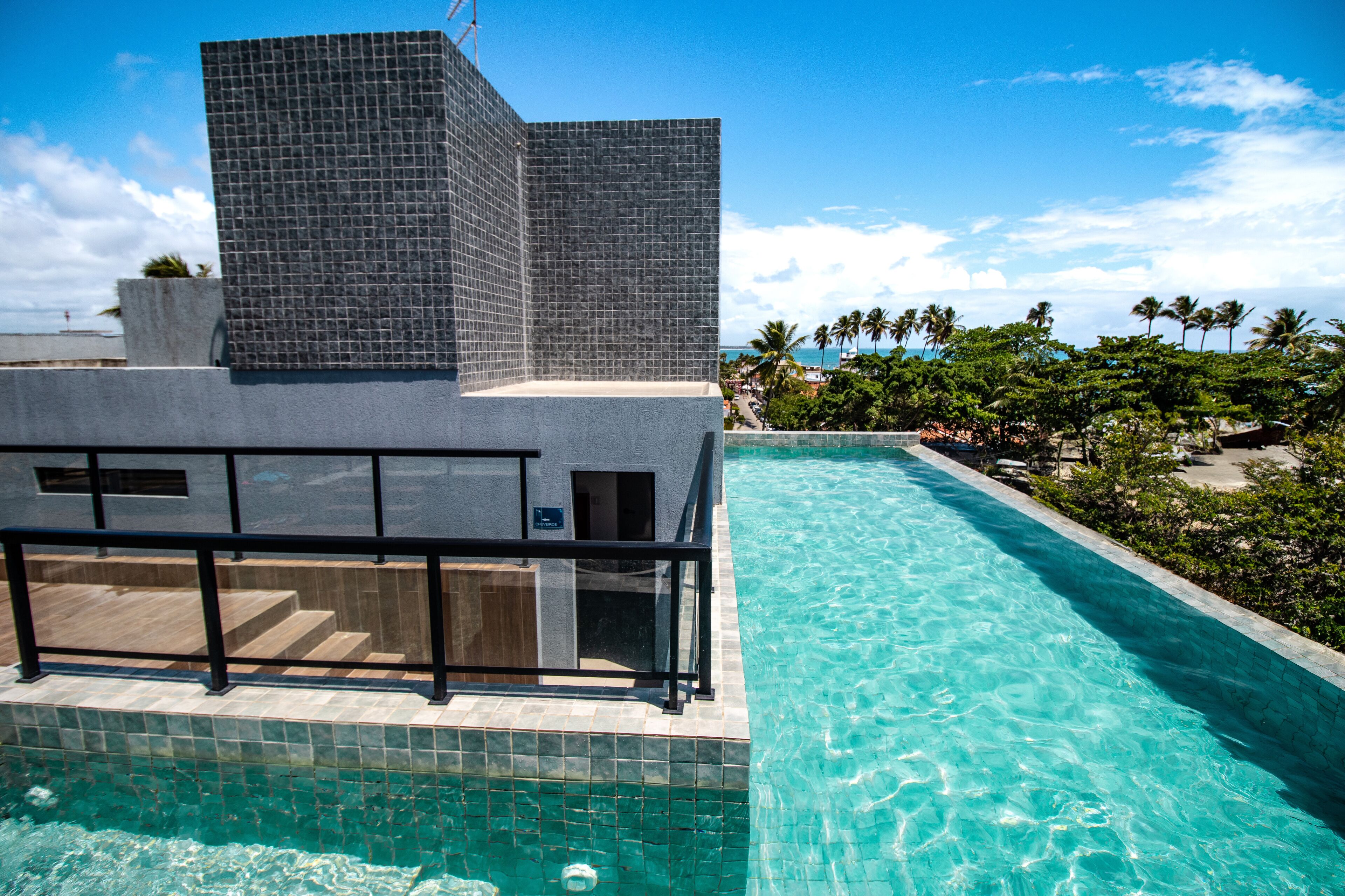 Una piscina con agua cristalina, un edificio moderno con fachada de azulejos y una vista de palmeras y una playa al fondo.