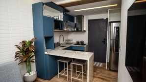 A compact kitchen with a built-in island, a grey sofa, and a potted plant. - Residencial Di Mare - by New Time  (Ipojuca)