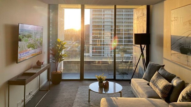 Una sala de estar moderna con un televisor grande, un sofá, una mesa de centro y un balcón con vistas a un paisaje urbano.