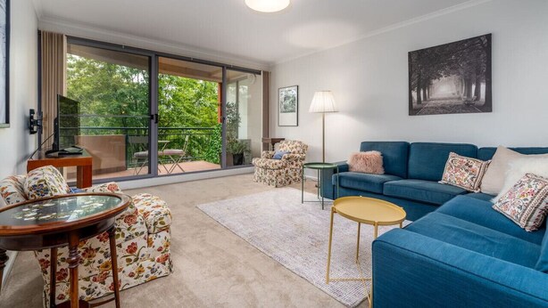 Una sala de estar moderna con un sofá azul, un sillón estampado y una mesa de centro de vidrio. Hay un gran ventanal corredera que da a un balcón con vistas a la vegetación.